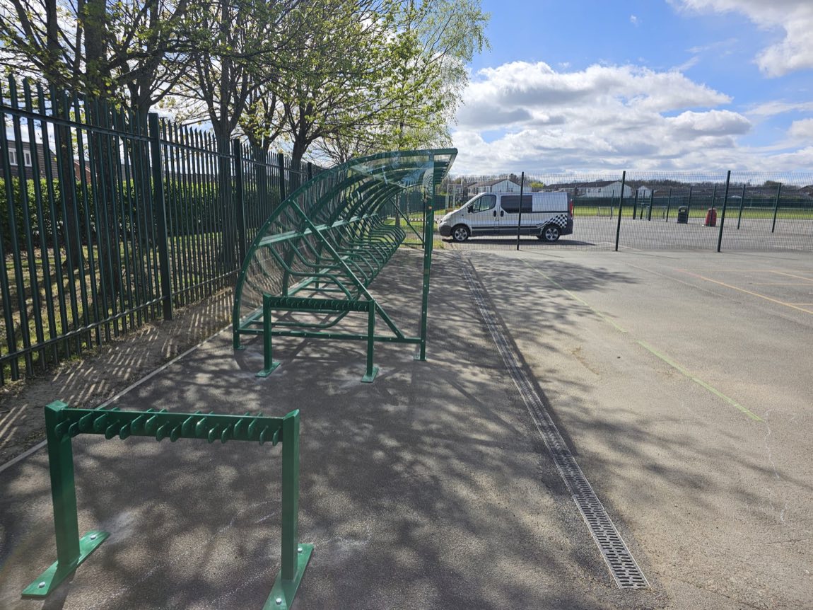 Green cycle shelter and separate rack beside a fence with a parked van