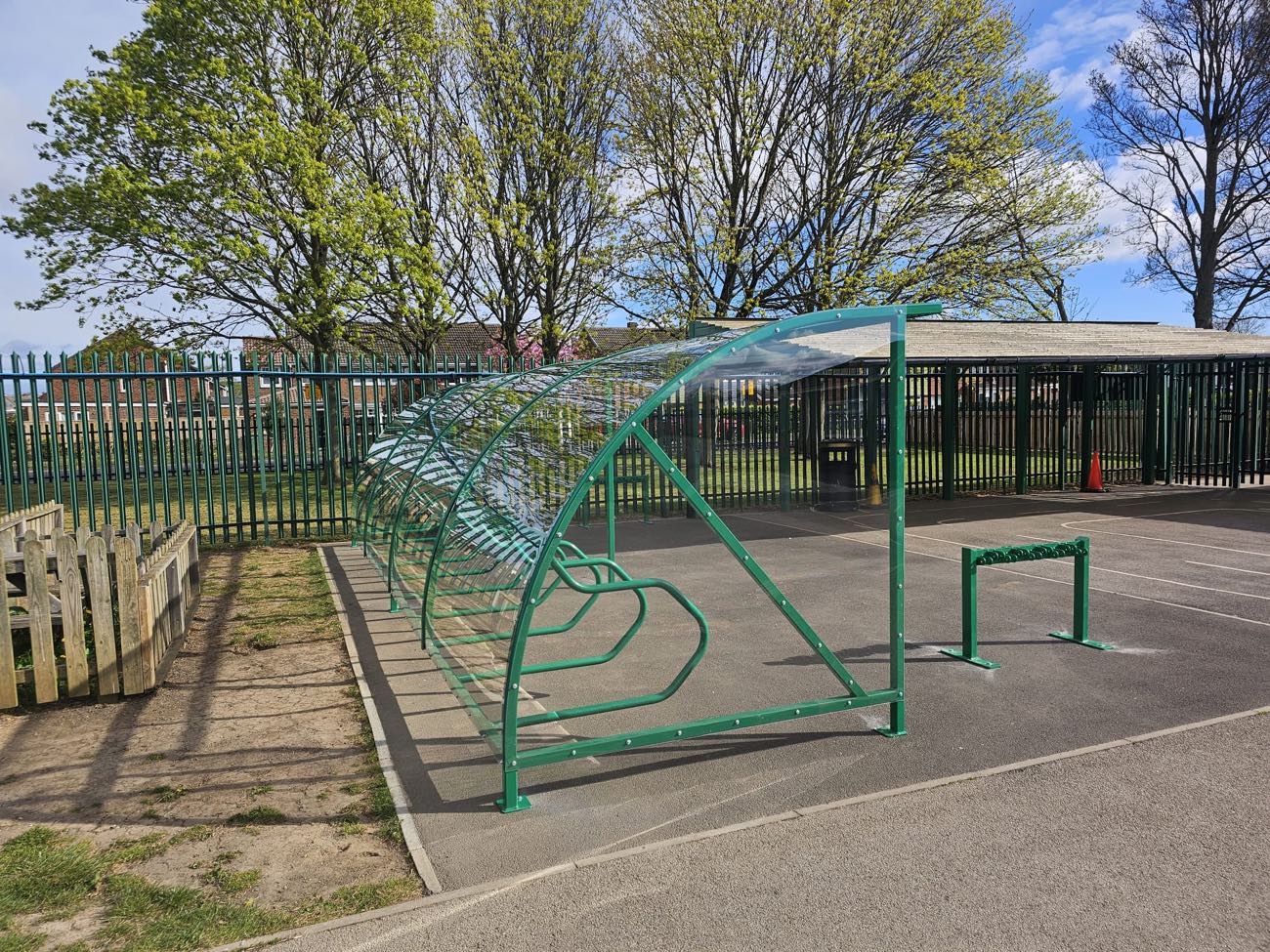 Green curved cycle shelter with cycle racks beside a school playground fence