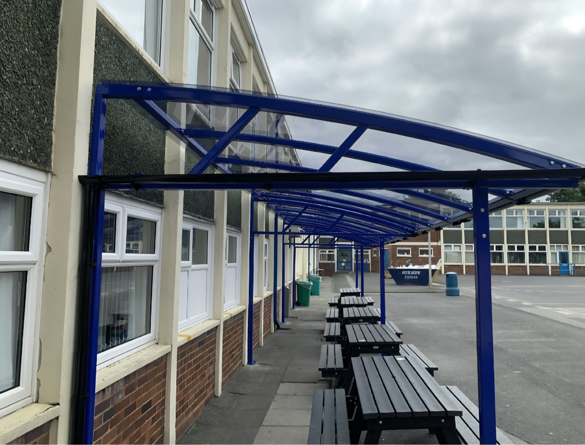Blue curved dining canopy with a clear polycarbonate roof over picnic benches along a school building in Bristol