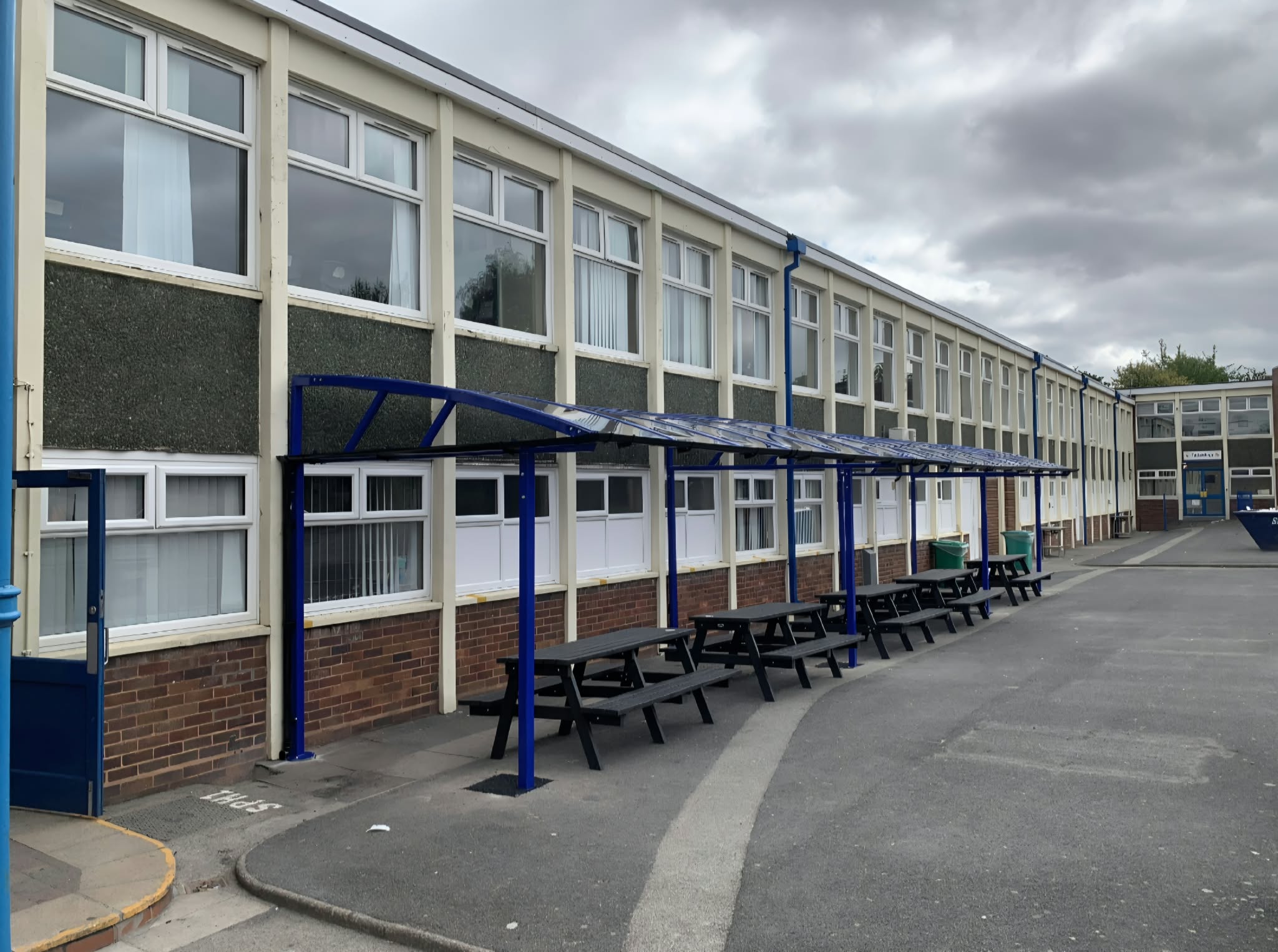 Side view along the blue dining canopy and its row of black picnic benches on a school playground