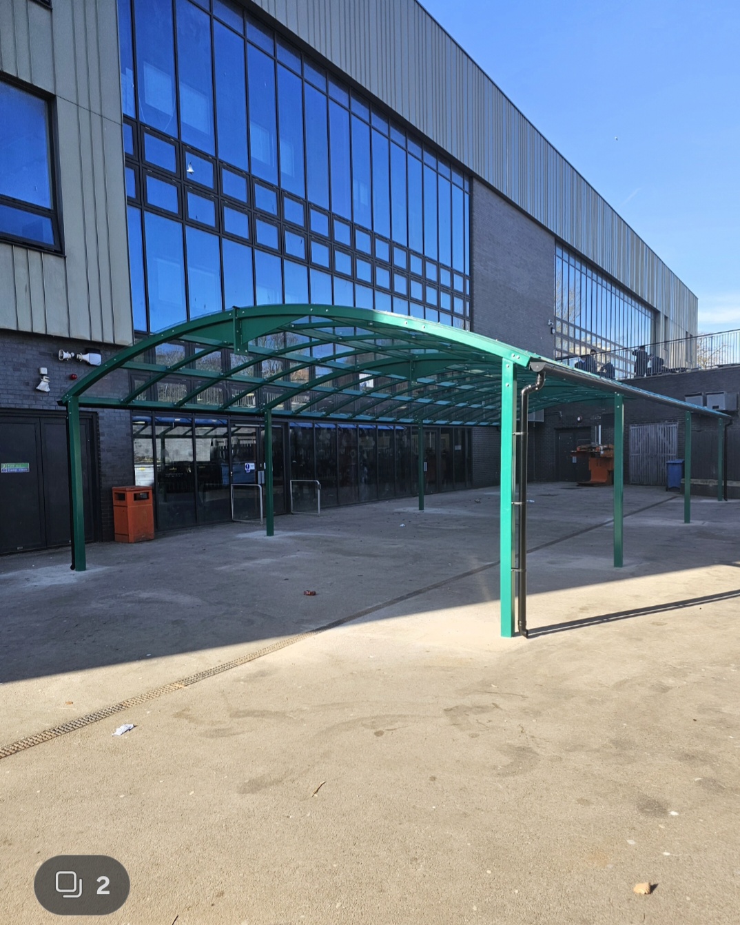 Green curved dining canopy with a clear polycarbonate roof on a paved yard outside a large glazed building in Chester