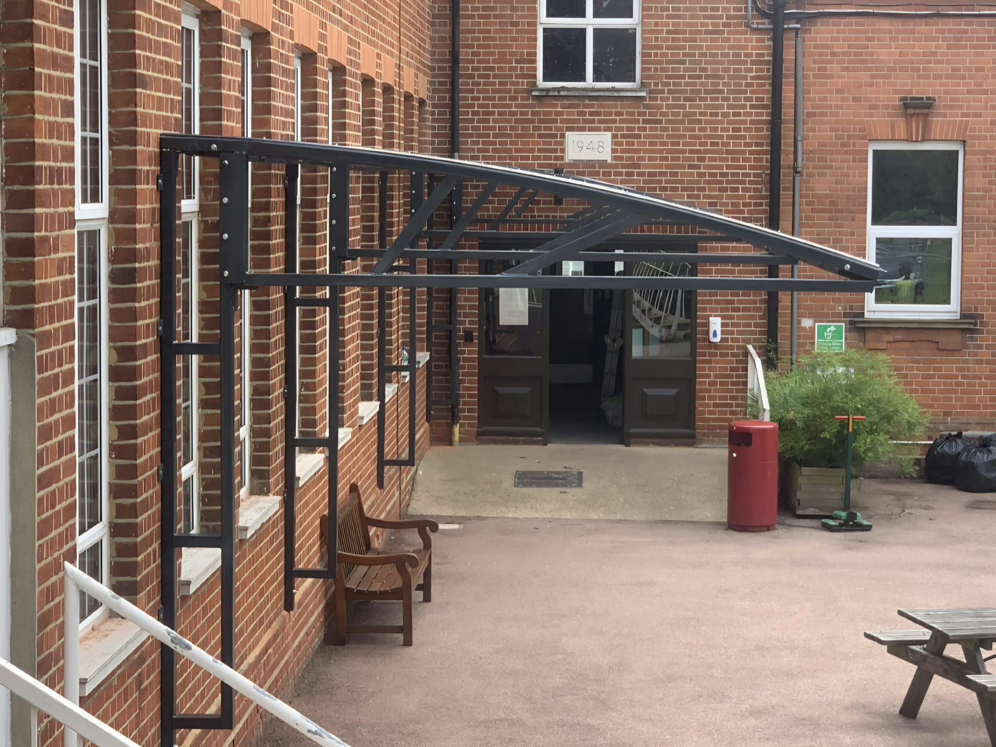 Side view of the anthracite canopy frame over a wooden bench against the red-brick building