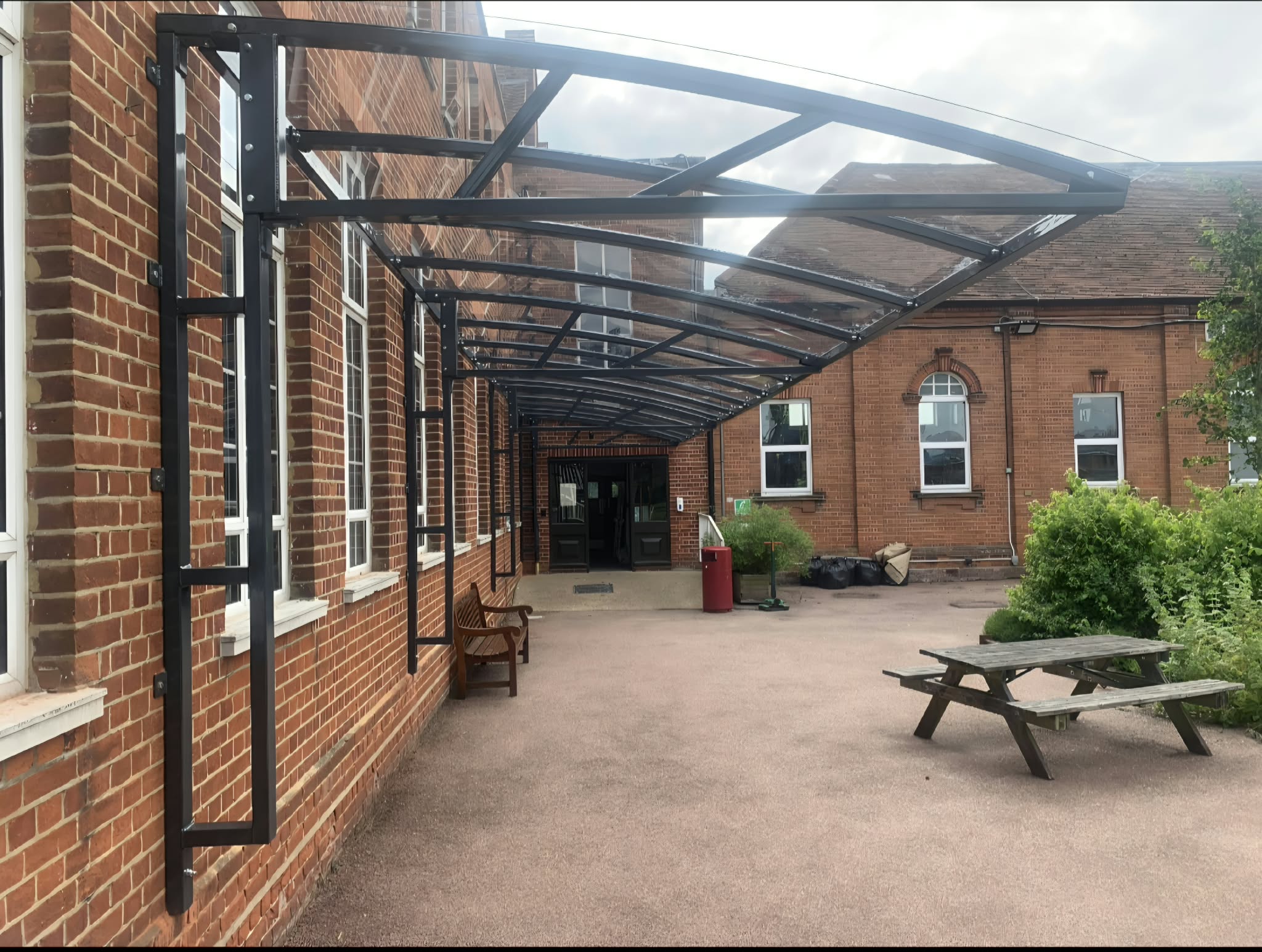 Looking along under the black canopy's clear curved roof to an entrance, with a picnic table on the courtyard