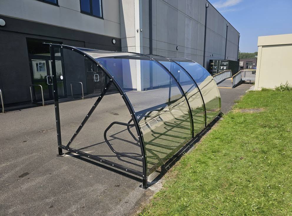 Black cycle shelter with clear curved roof and cycle rack beside the building at Ellesmere Park High School, Manchester
