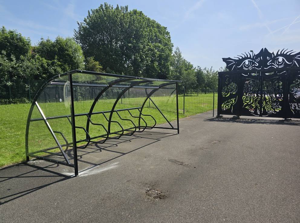 Open-sided cycle shelter frame on tarmac beside a grass field and a decorative gated entrance