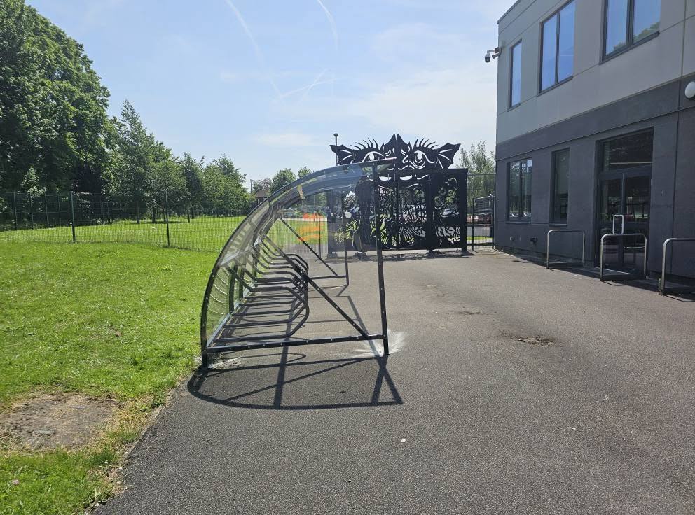Cycle shelter end-on with its curved clear roof, set on a tarmac path by the grounds at Ellesmere Park High School