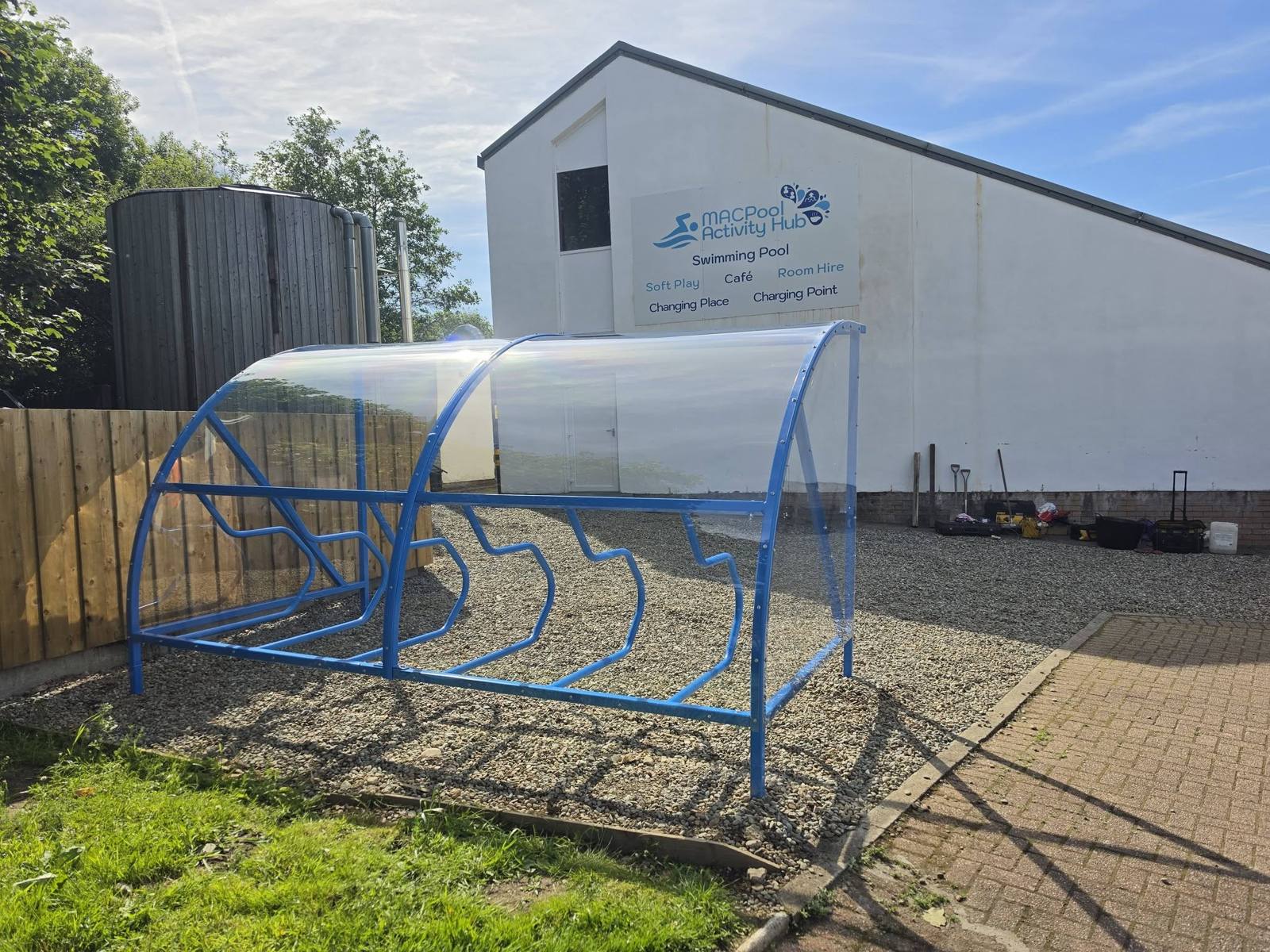 Side view of the blue cycle shelter on gravel, with the MACPool Activity Hub swimming pool signage behind