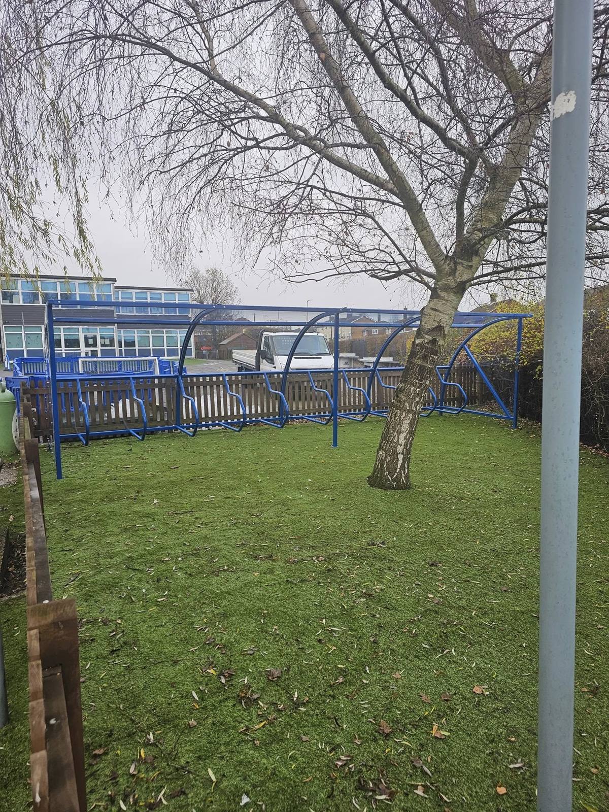 Long blue cycle shelter running along a wooden fence on artificial grass, a bare tree and school building behind