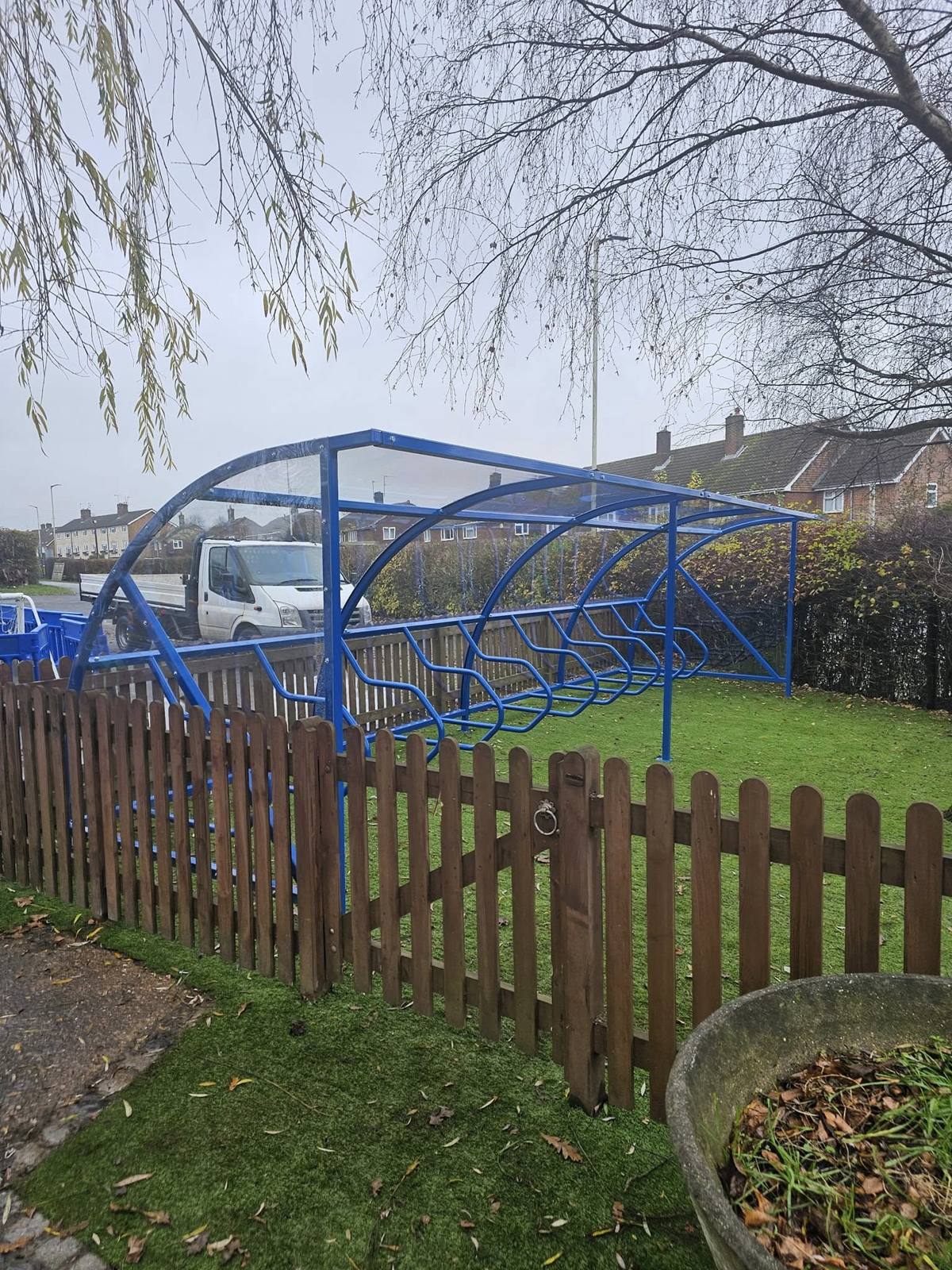 Blue cycle shelter with clear sloping roof behind a picket fence, houses and a flatbed van across the road