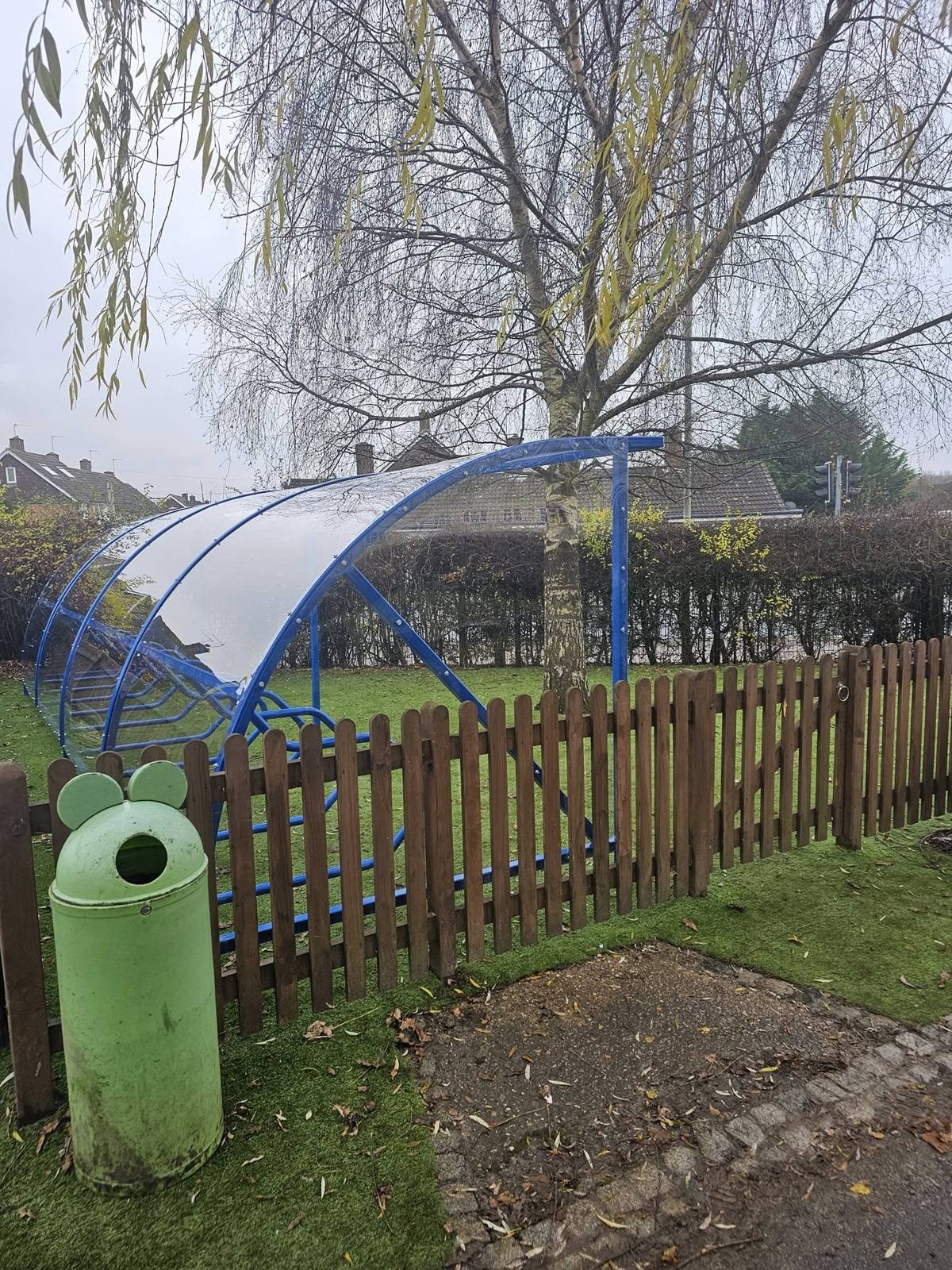 Blue cycle shelter seen end-on behind a wooden fence, a mouse-eared green litter bin in the foreground