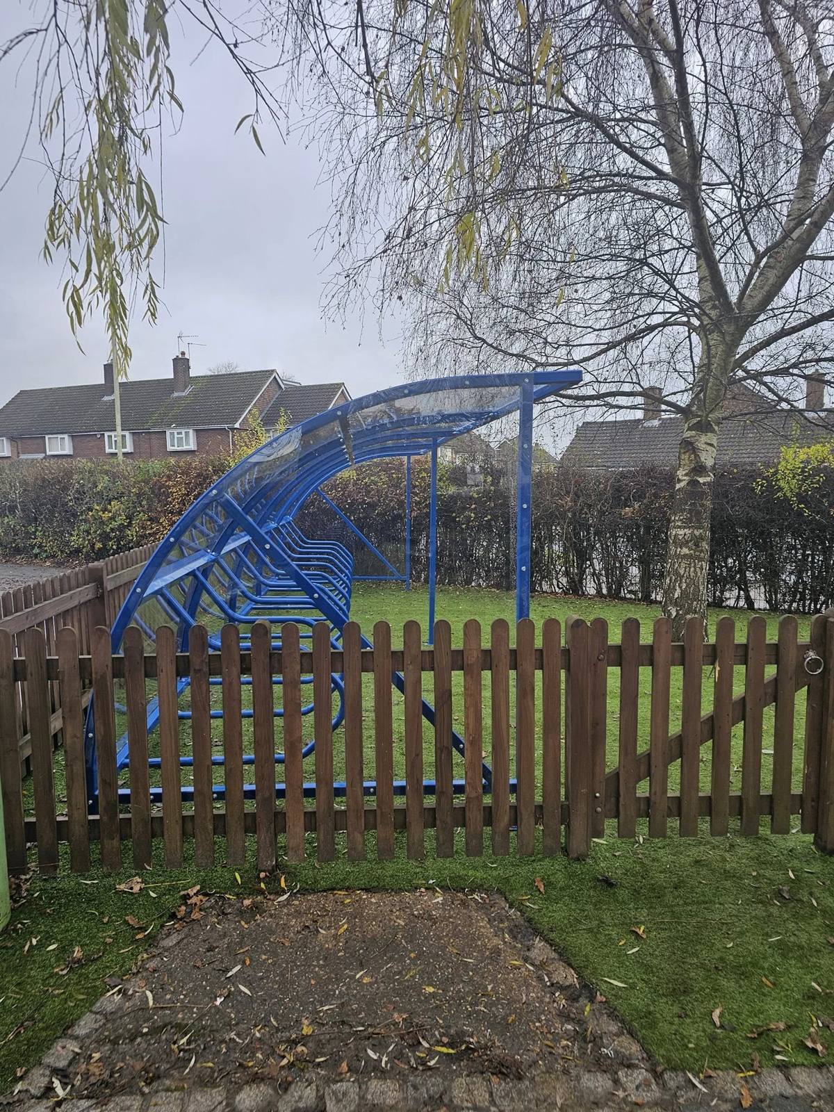 End view of a blue cycle shelter with curved clear roof behind a picket fence at Micklem Primary School, Hemel Hempstead