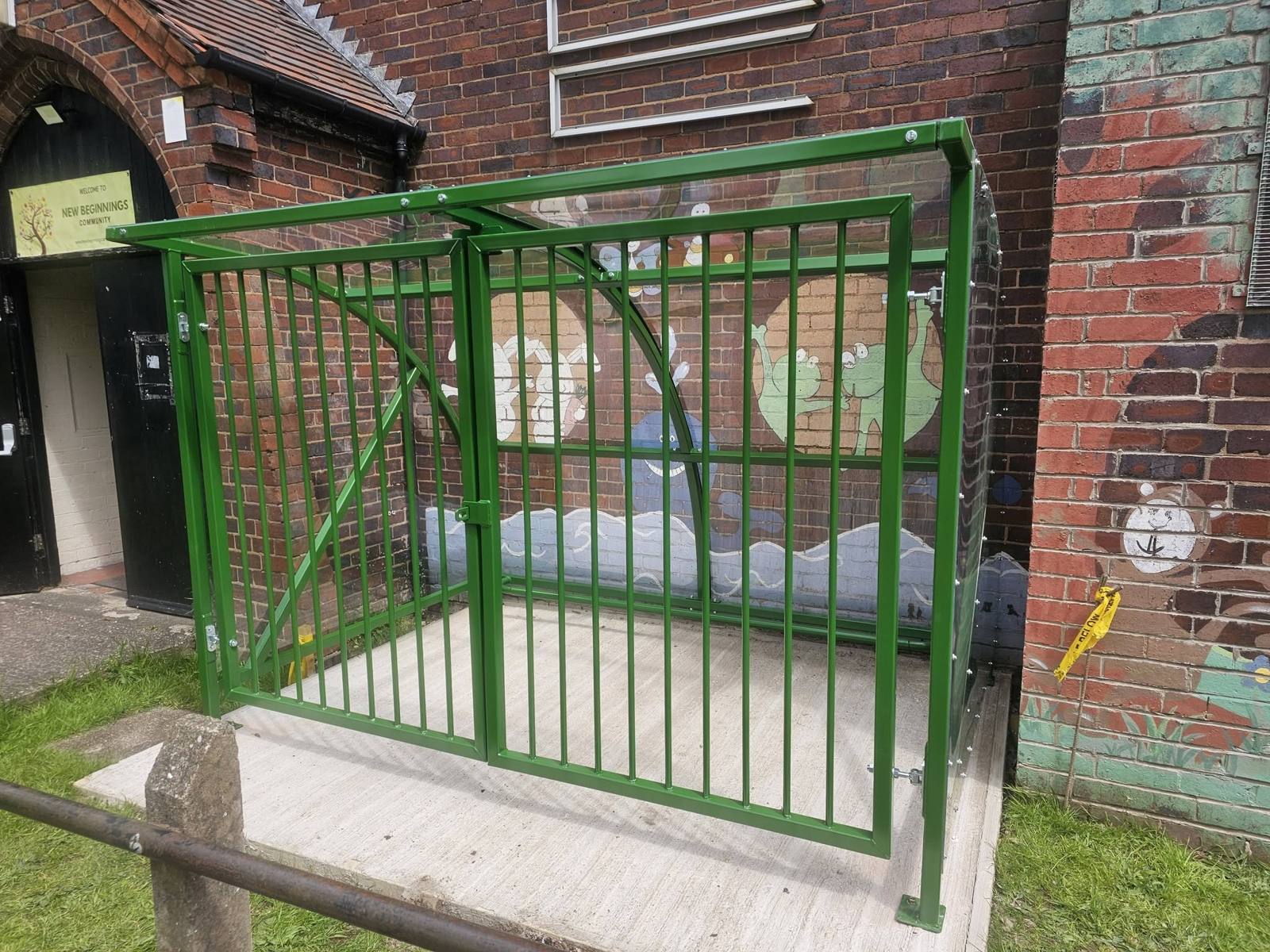 Green buggy shelter with barred security gates against a painted brick wall at New Beginnings Creative, Birmingham