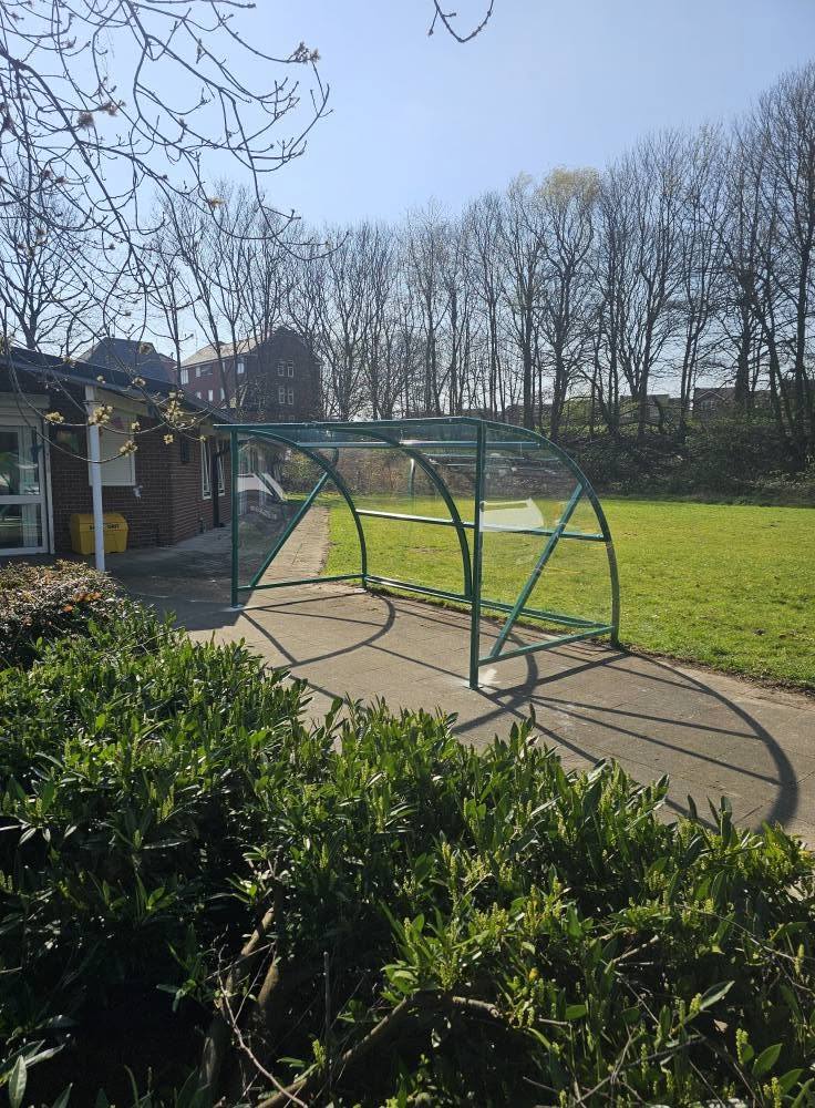 Distant view of the green curved shelter on a paved path with grass and trees behind