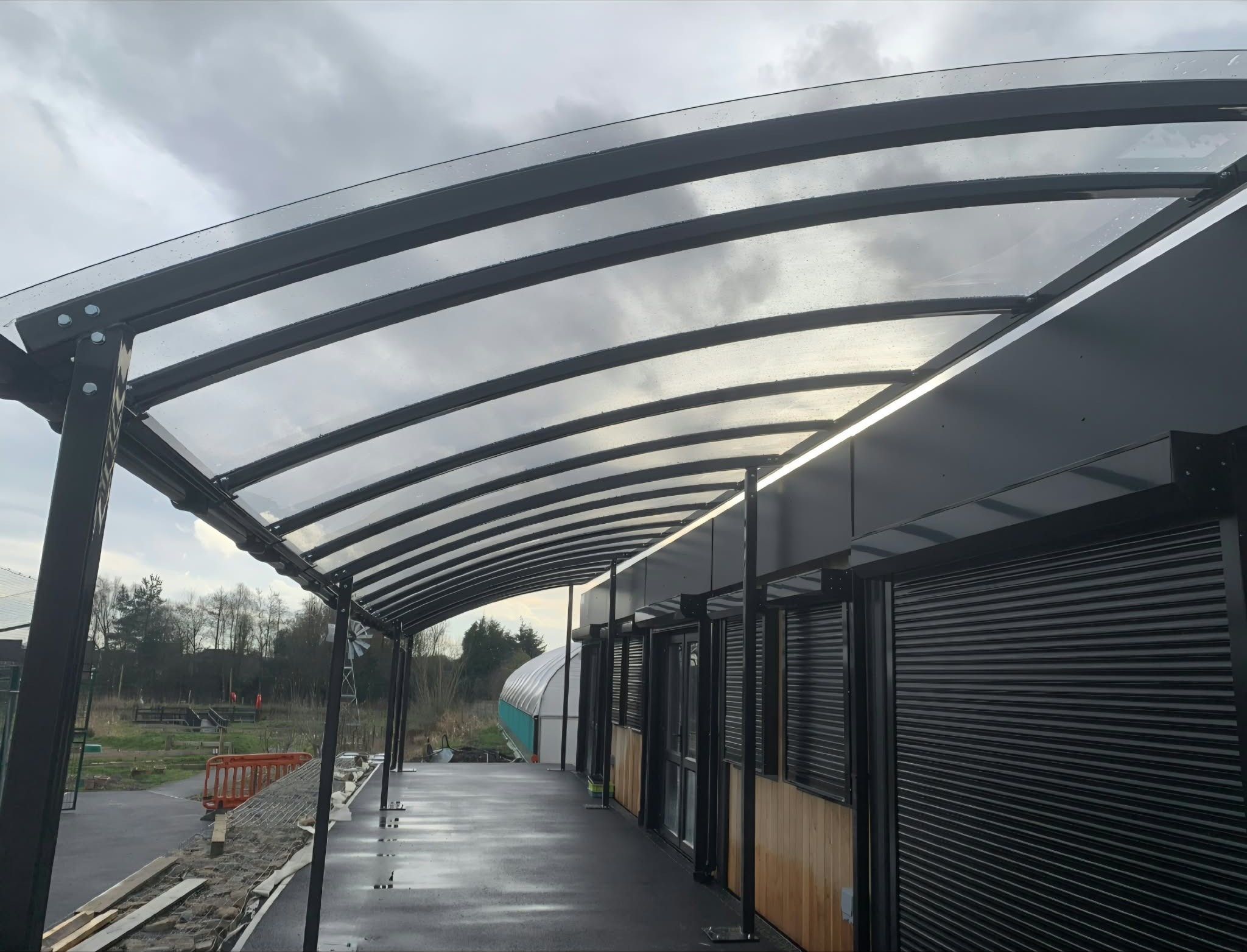 Under the curved canopy looking along the covered walkway beside the school building's shutters and glazing