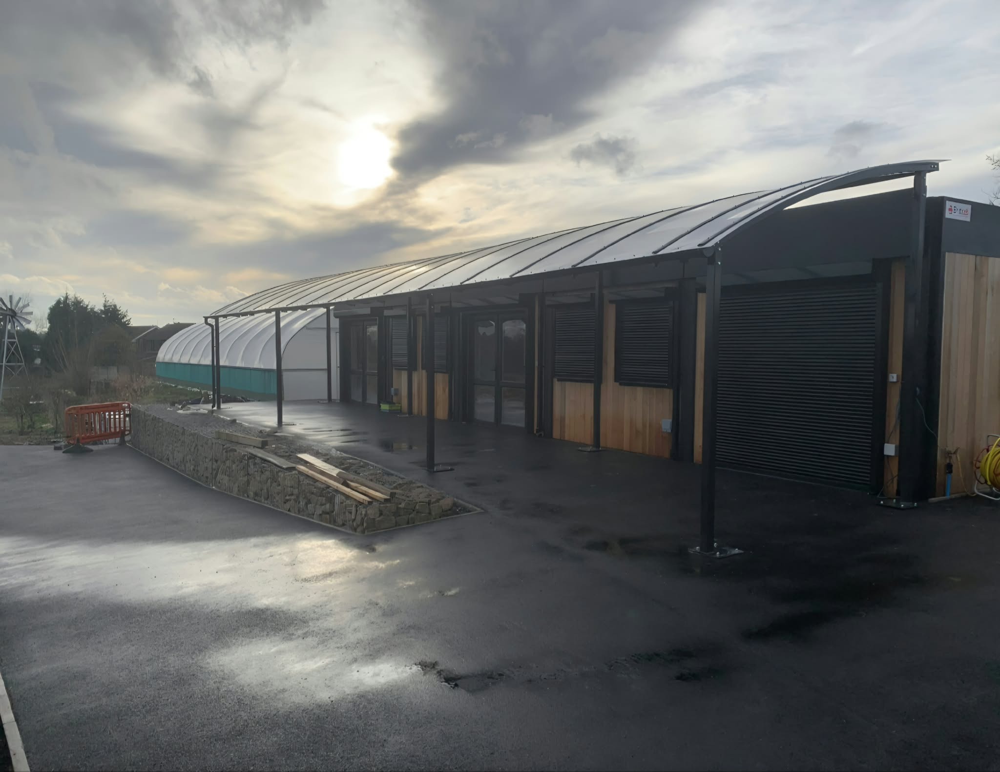 Wide view of the dark curved canopy running the length of the modular school building beside a polytunnel