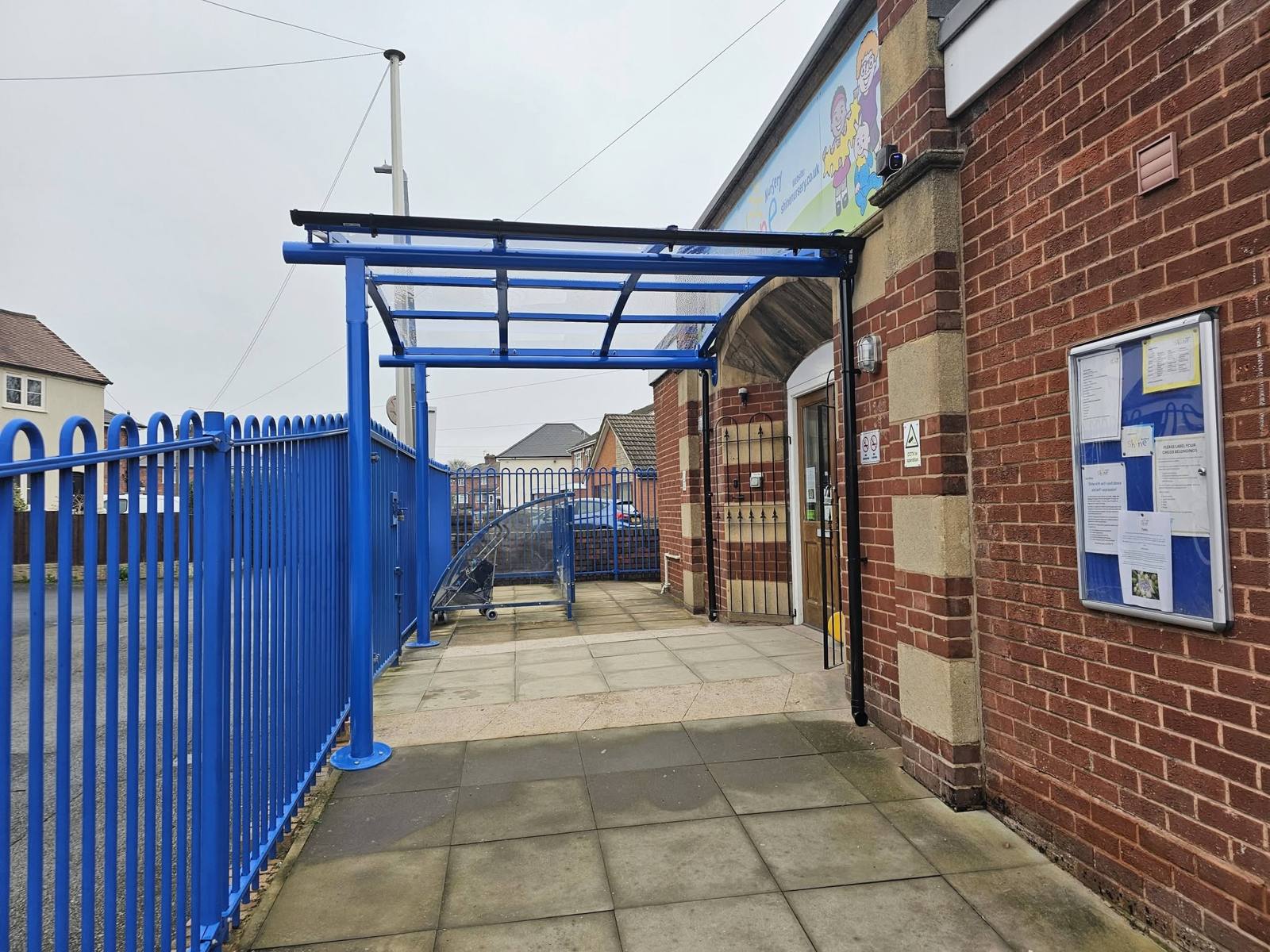 Underside view of the blue entrance canopy along the paved waiting area beside the railings