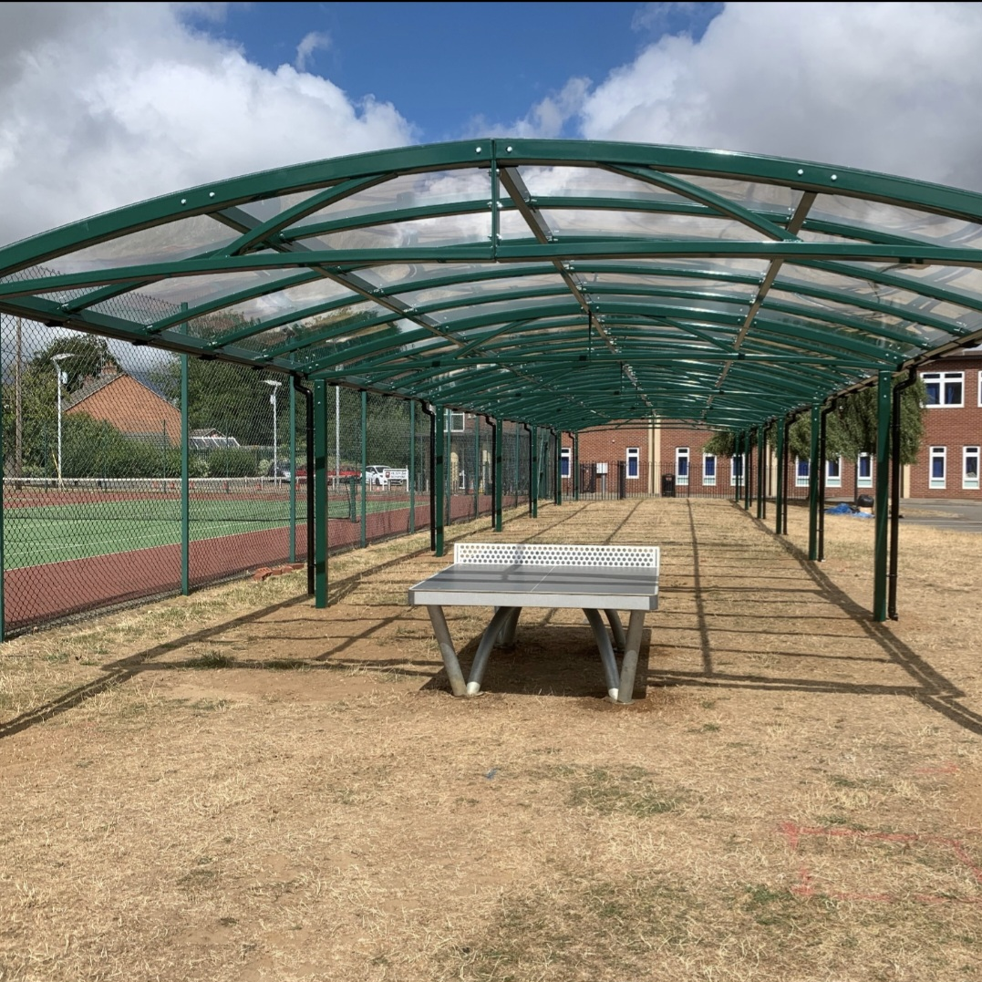 Green barrel-vaulted sports canopy with a clear polycarbonate roof beside a fenced MUGA and a table-tennis table, in Leicester