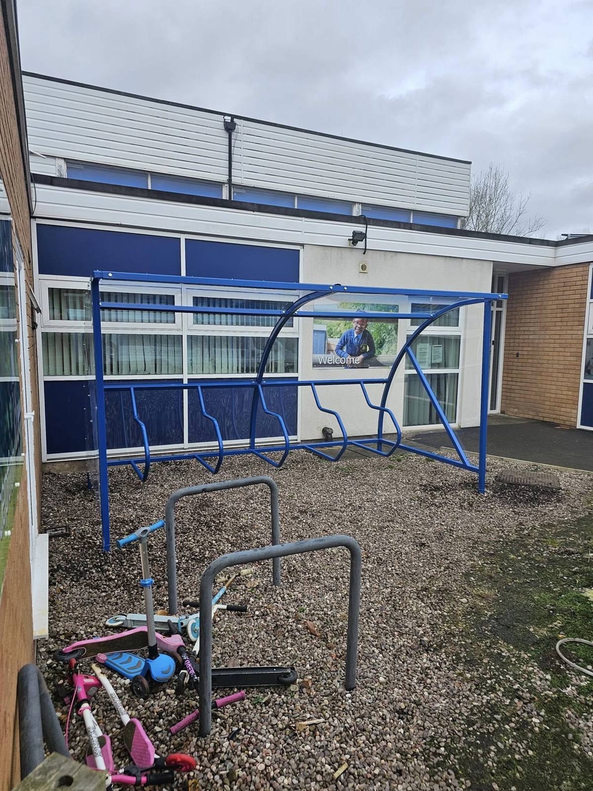 Blue steel cycle shelter with clear curved roof against a school building with a Welcome sign, scooters scattered on gravel in front
