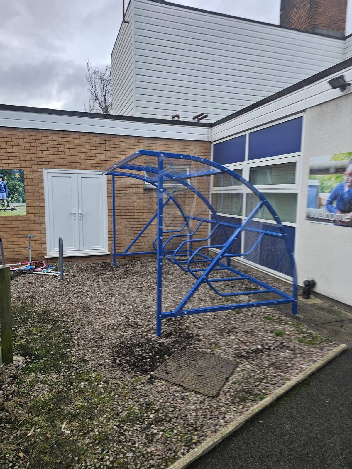 End-on view of a blue cycle shelter showing its curved frame and tiered rack beside a brick wall and white doors