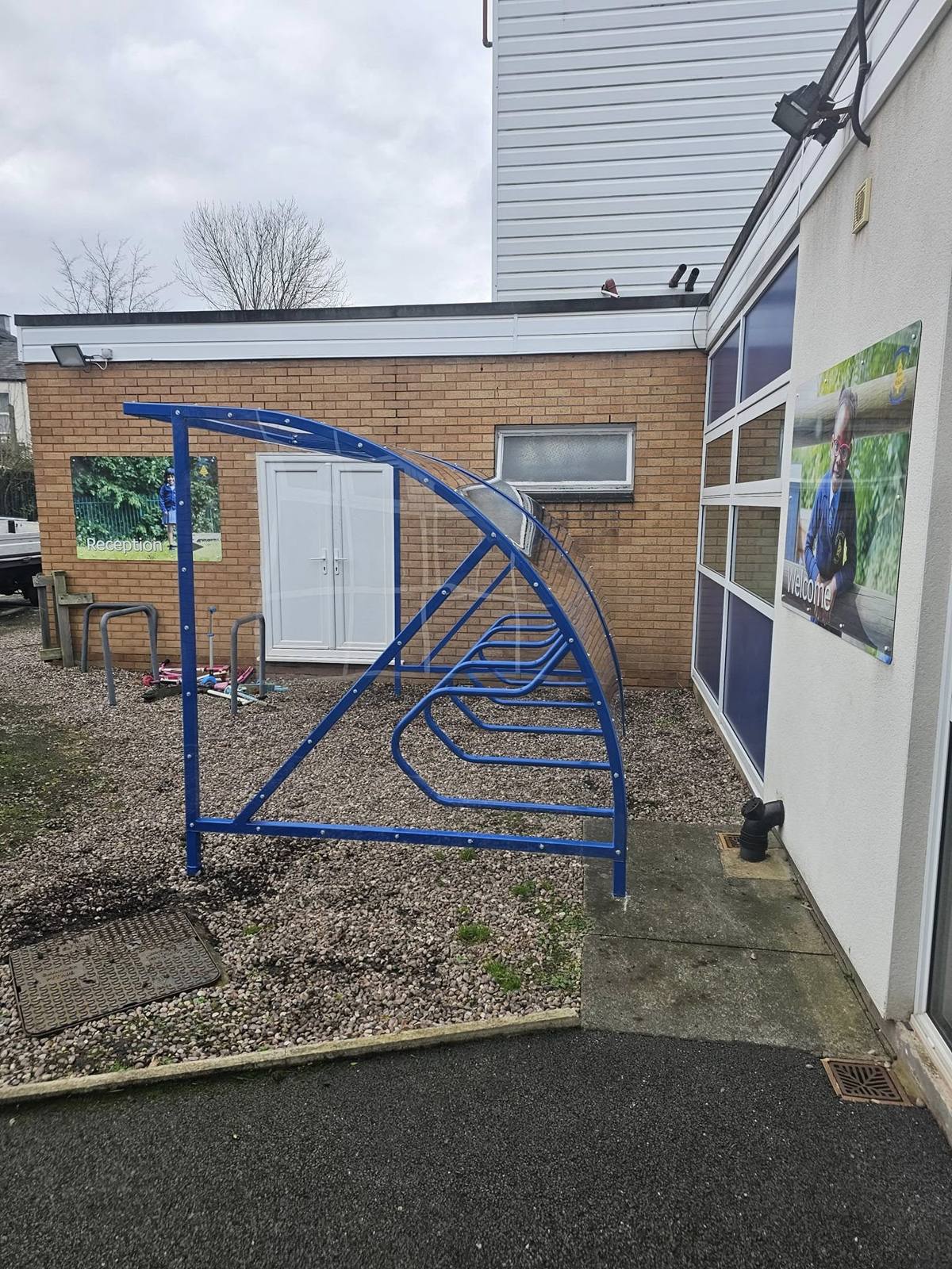 Angled view of a blue cycle shelter on gravel between a brick wall and a window bearing a Reception panel
