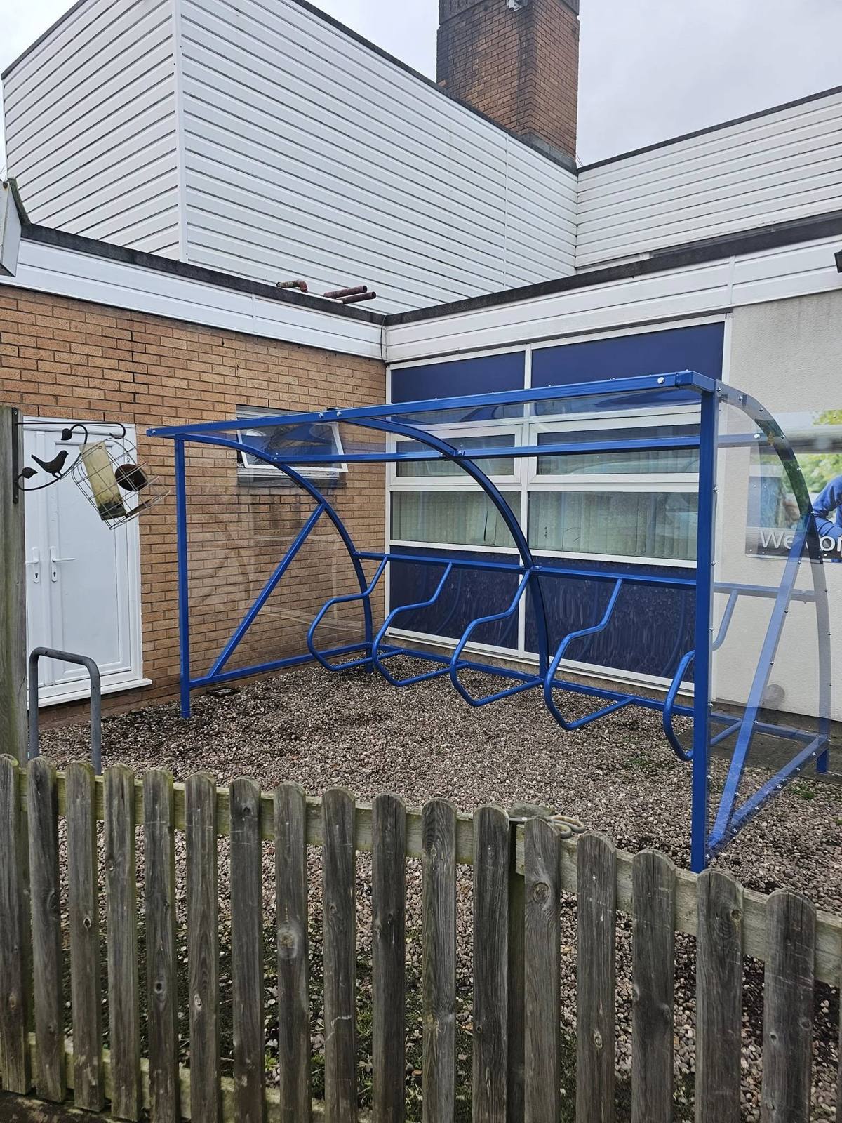 Blue cycle shelter with transparent roof seen from behind a wooden fence at St Boniface RC Primary School