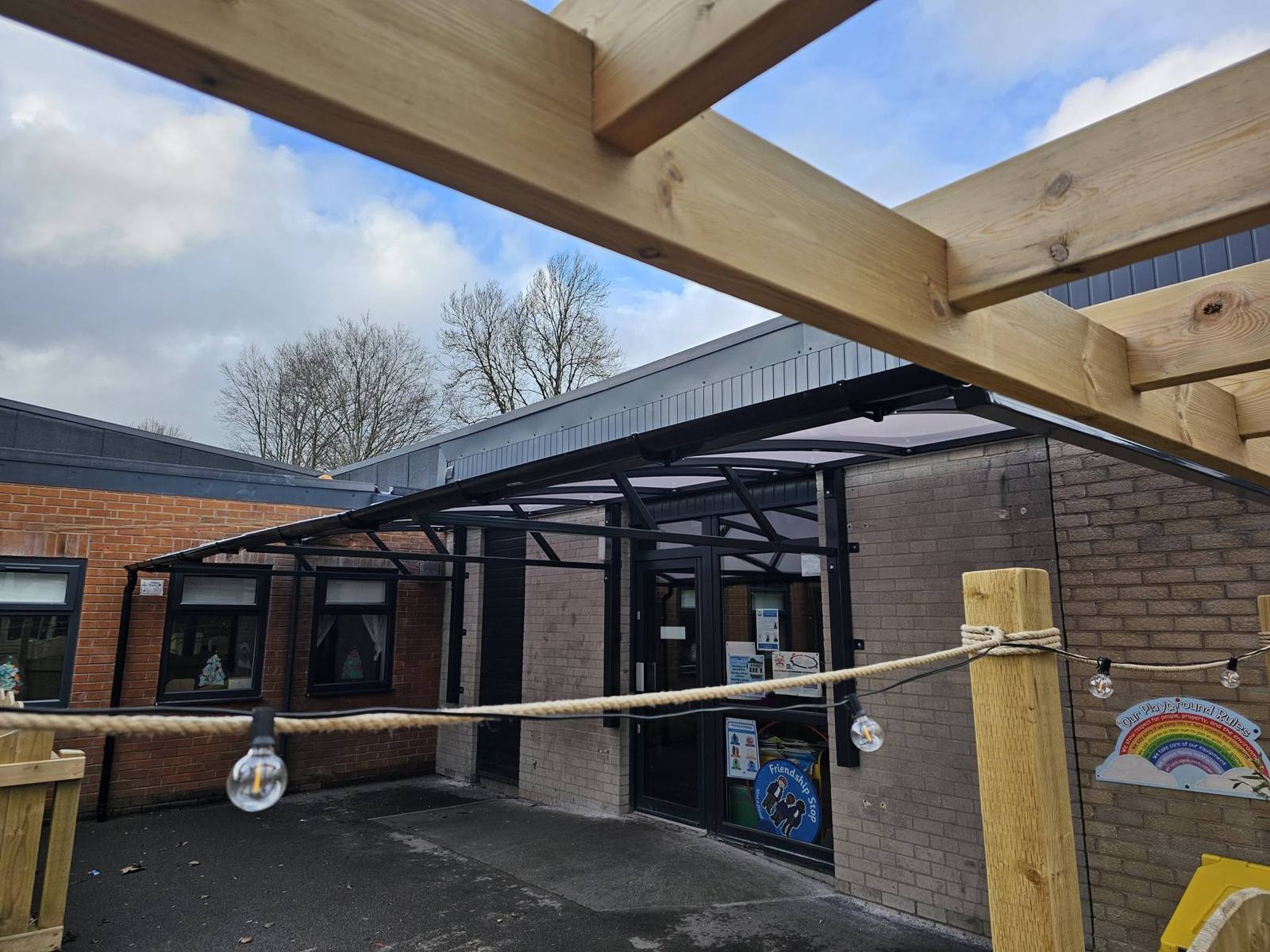 Angled view of a dark curved canopy beside a timber pergola in a playground at St Luke's CE Primary School, Heywood