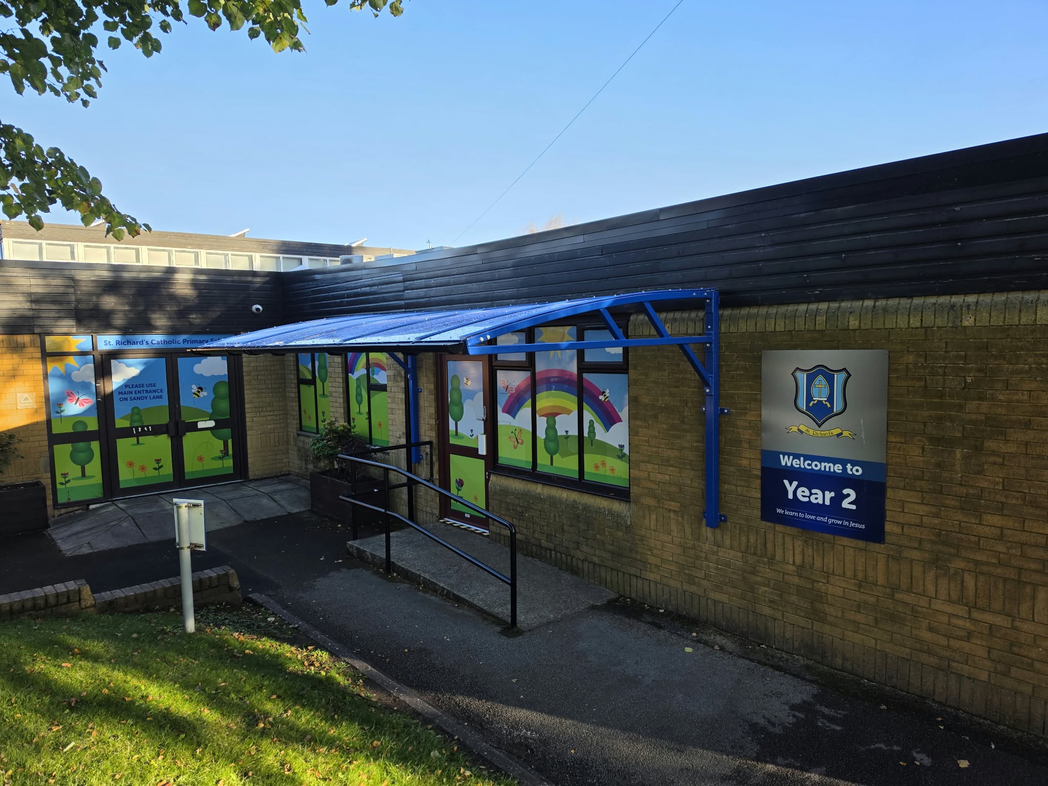 The blue wall-fixed cantilever waiting shelter and its clear roof over the ramped school entrance