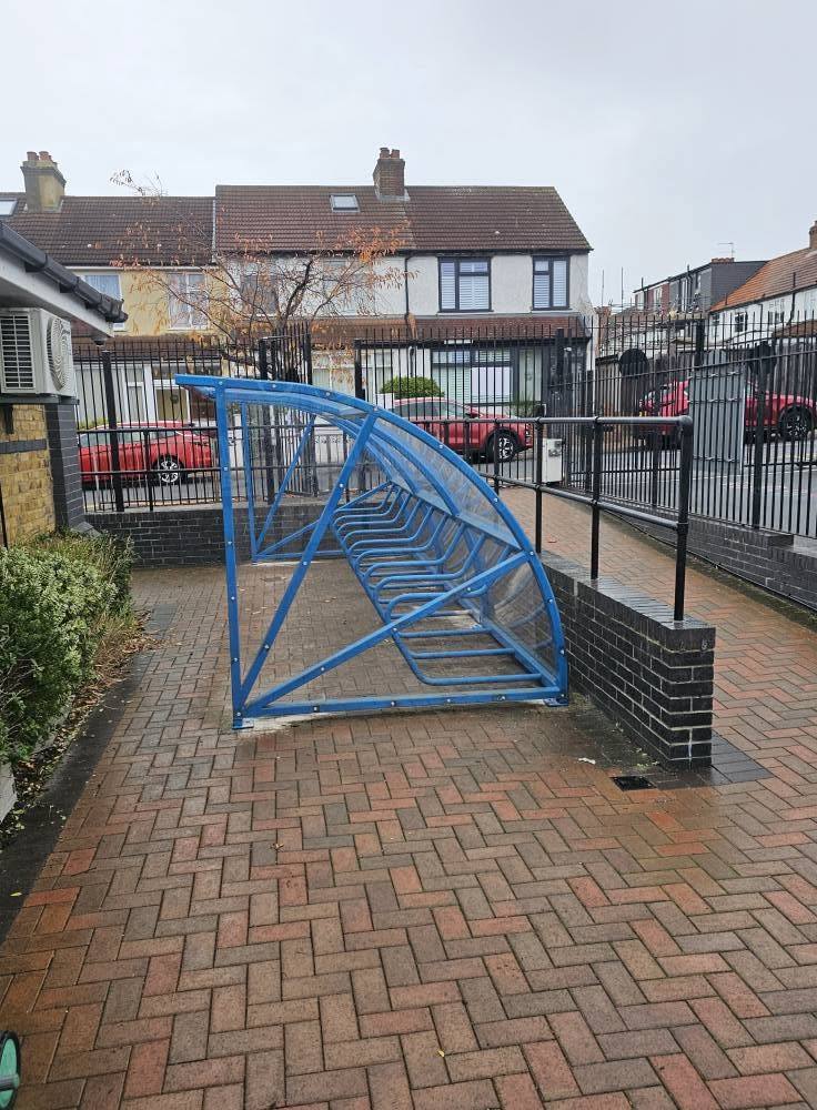 Blue cycle shelter with cycle stands inside, on block paving with houses behind