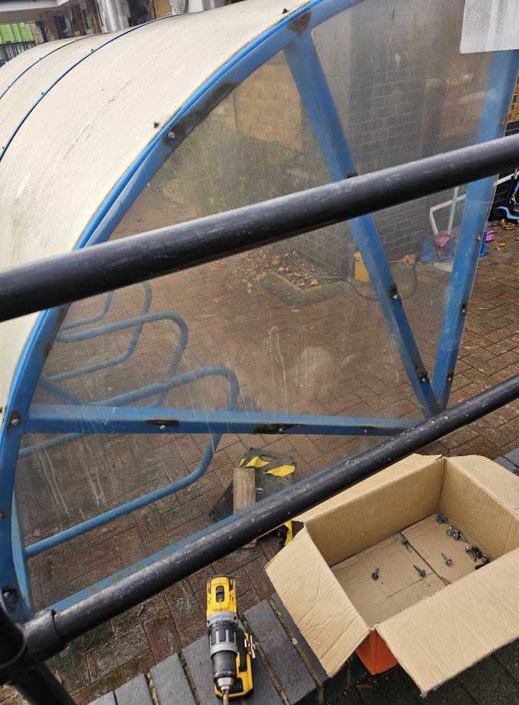Close view through the shelter's clear end panel to the cycle stands, with a drill and screws box below