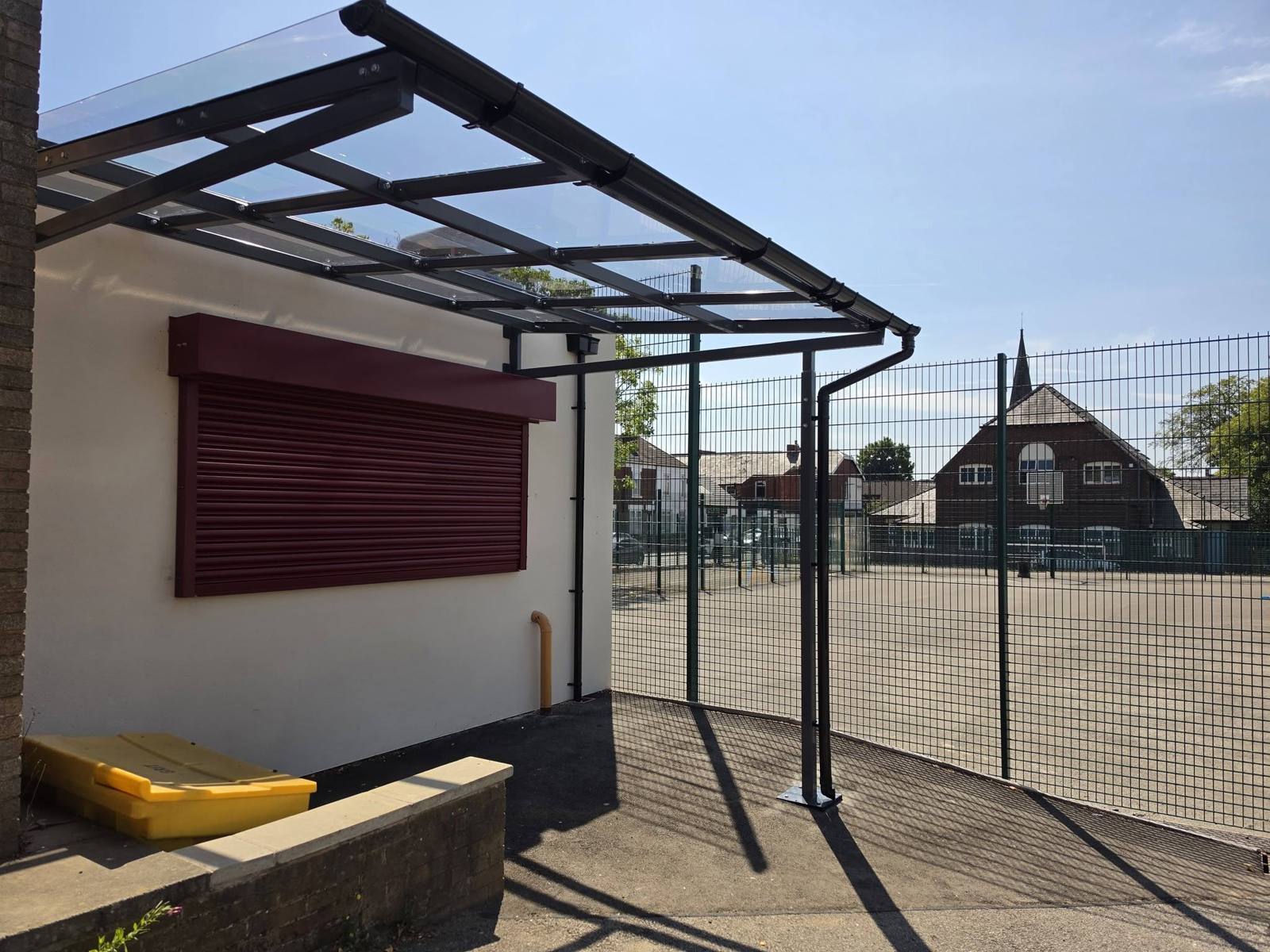 Black wall-mounted dining canopy with a clear glazed roof beside a maroon serving shutter at Stockport School