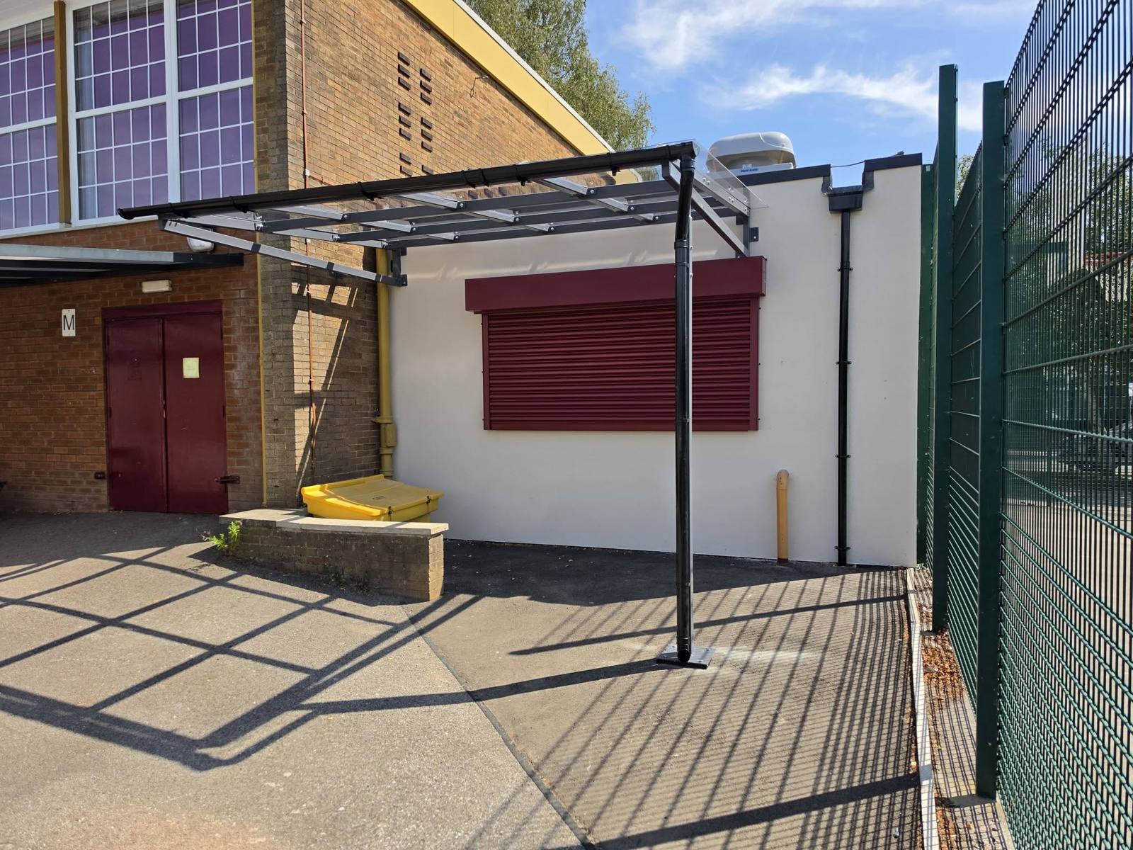 Dining canopy extending over the playground, sheltering a maroon roller shutter on a white servery wall