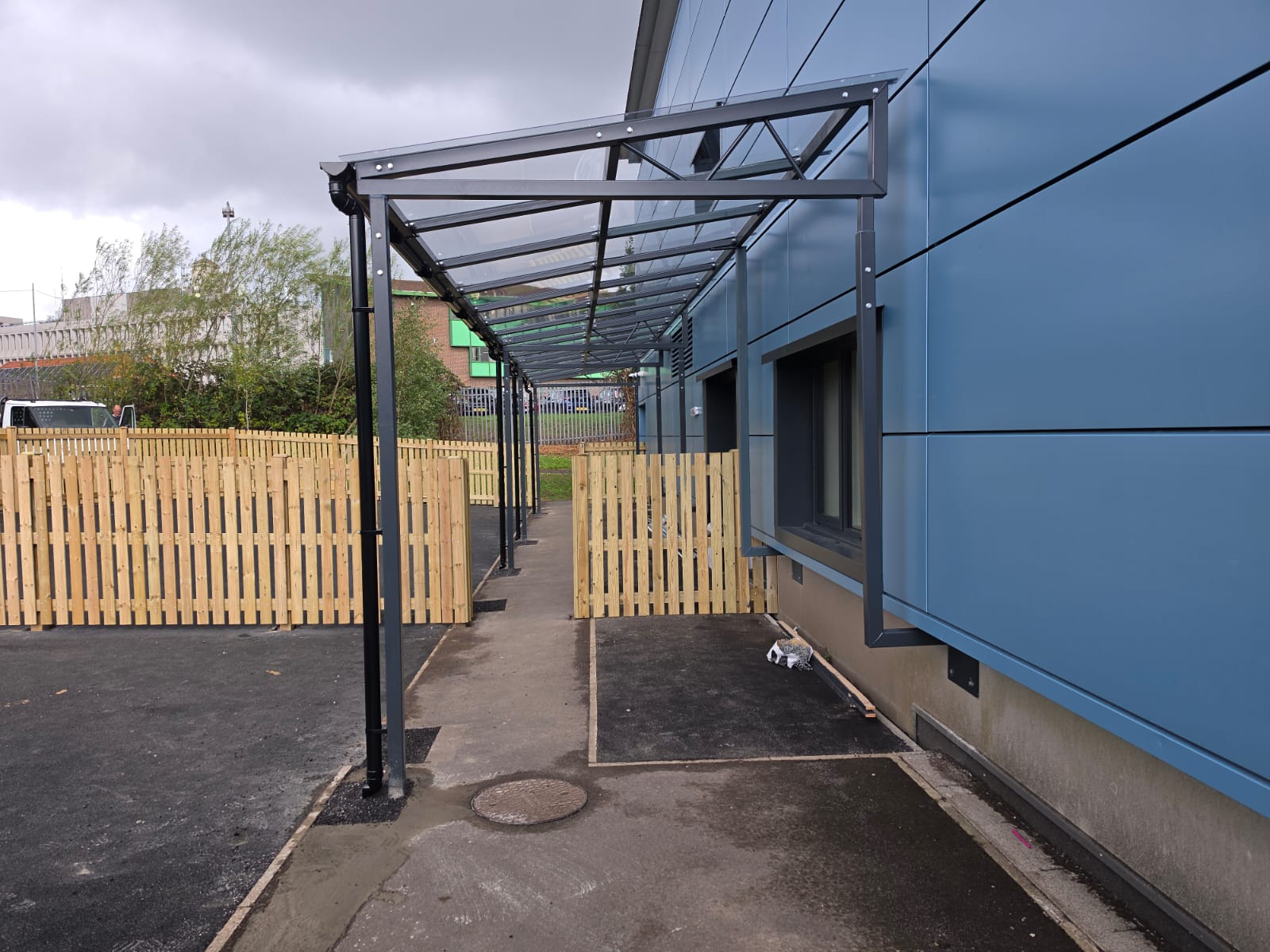 Walkway canopy sheltering a path between timber fencing and the blue cladding of The Deans School