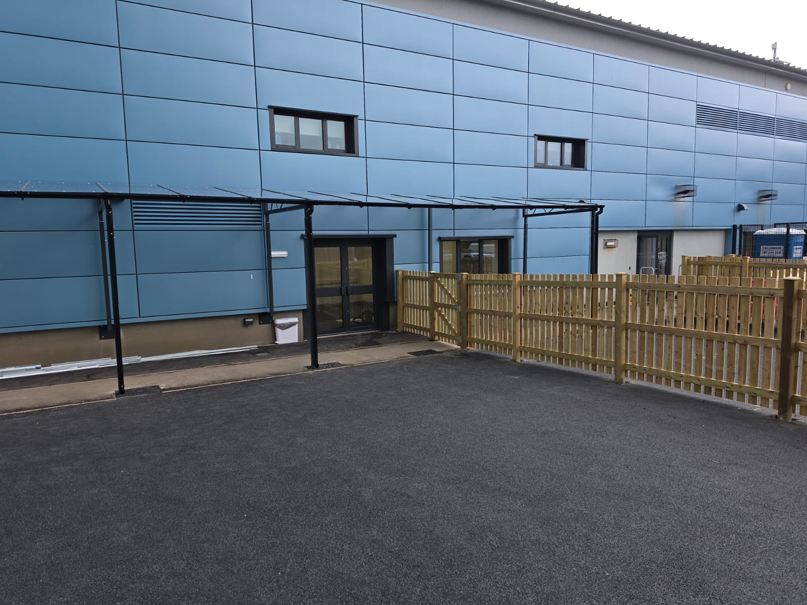 Long sloped canopy running the length of the blue-panelled building above doorways and a tarmac yard