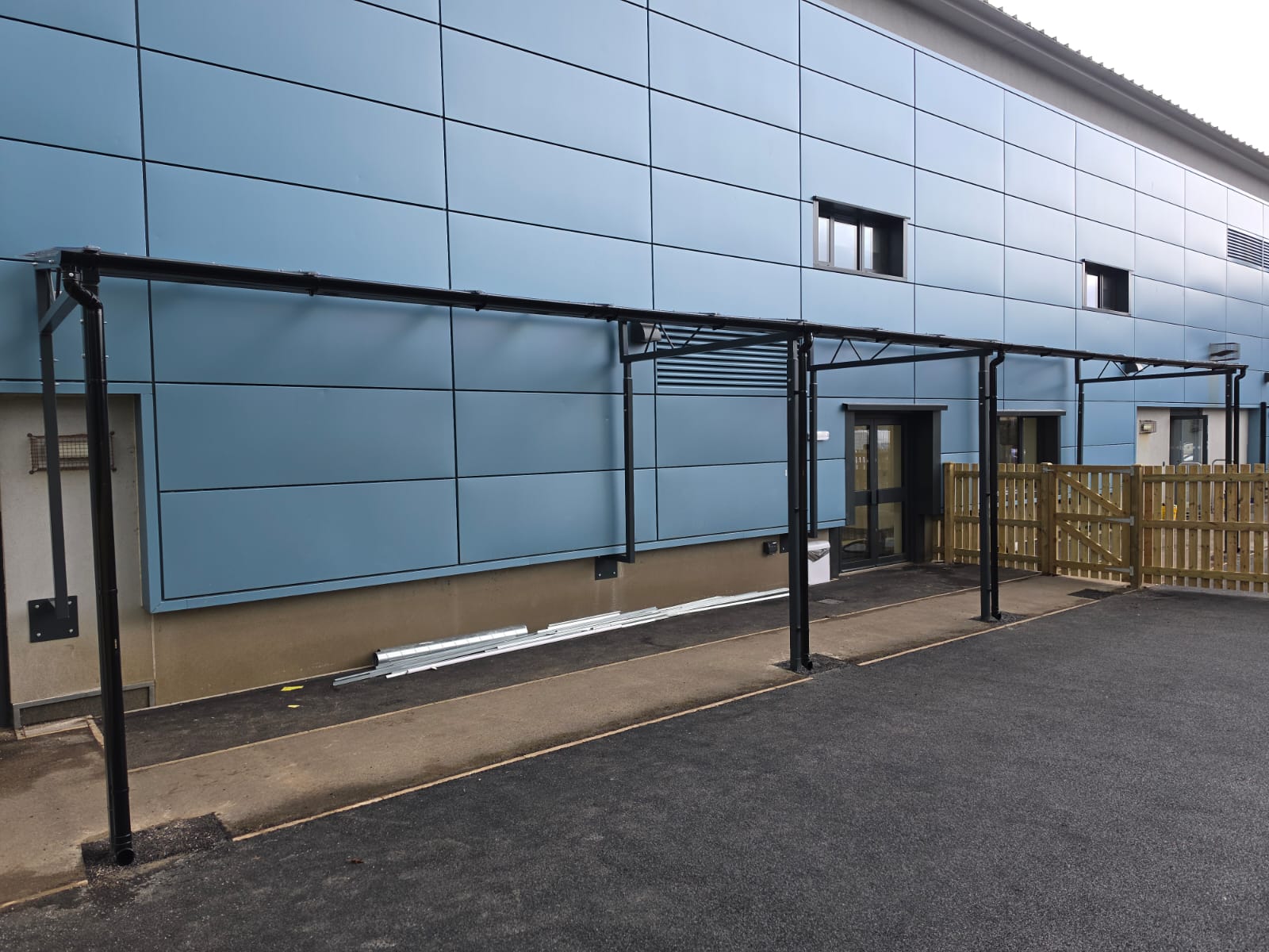Steel-framed canopy awaiting roof panels, spanning entrance doors along the blue-clad school wall