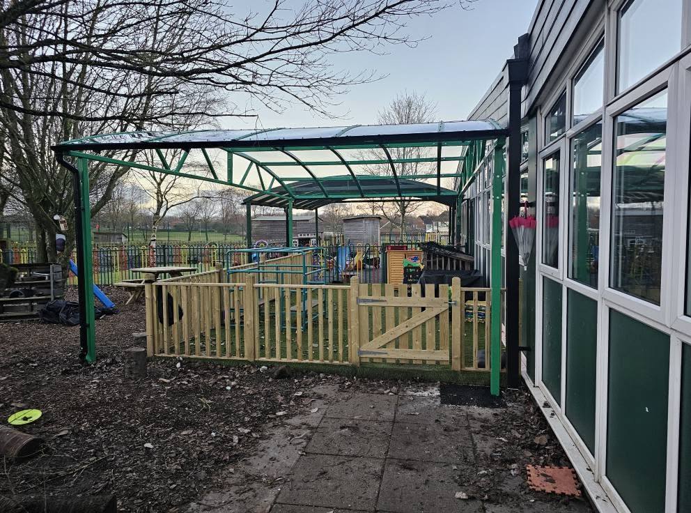 Green curved play-area canopy over a fenced playground beside the school building at Trinity Primary School