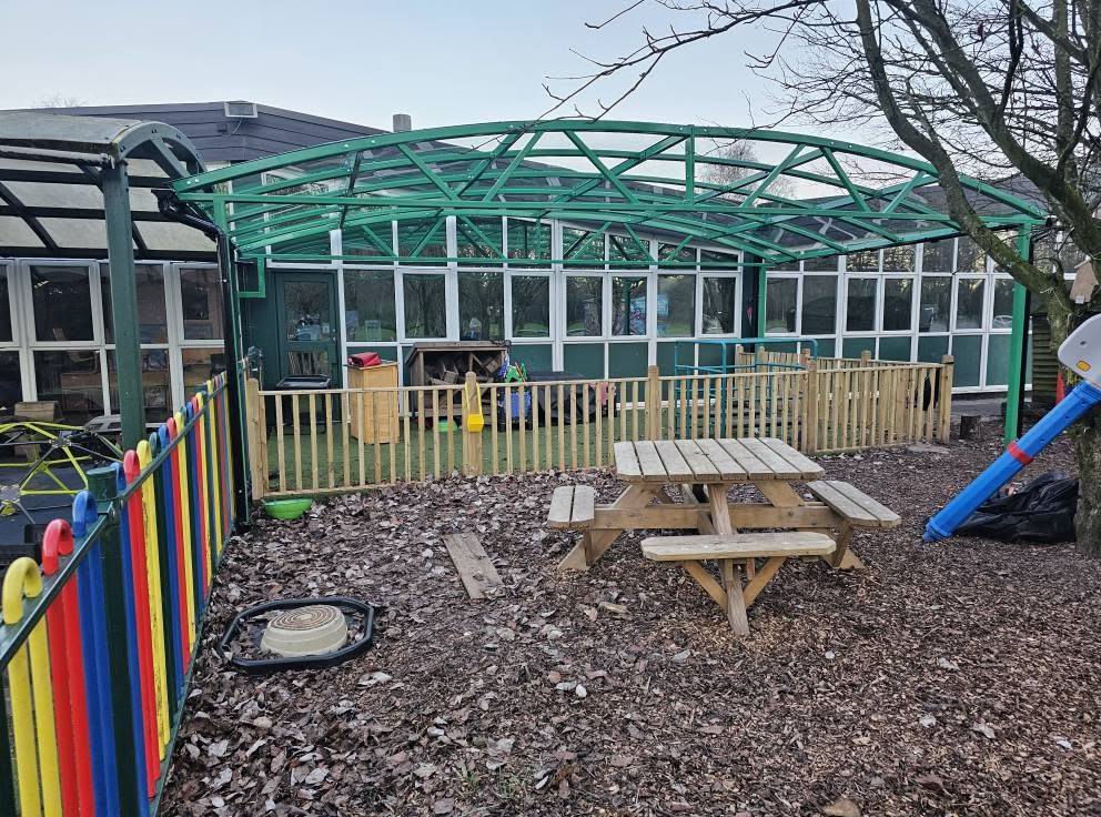 Green dome-framed play canopy alongside the school windows with a picnic bench on bark chippings