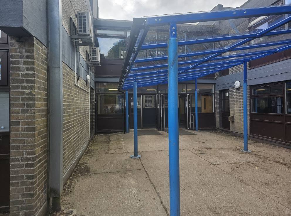 View along the blue-framed canopy leading towards glazed entrance doors in the courtyard