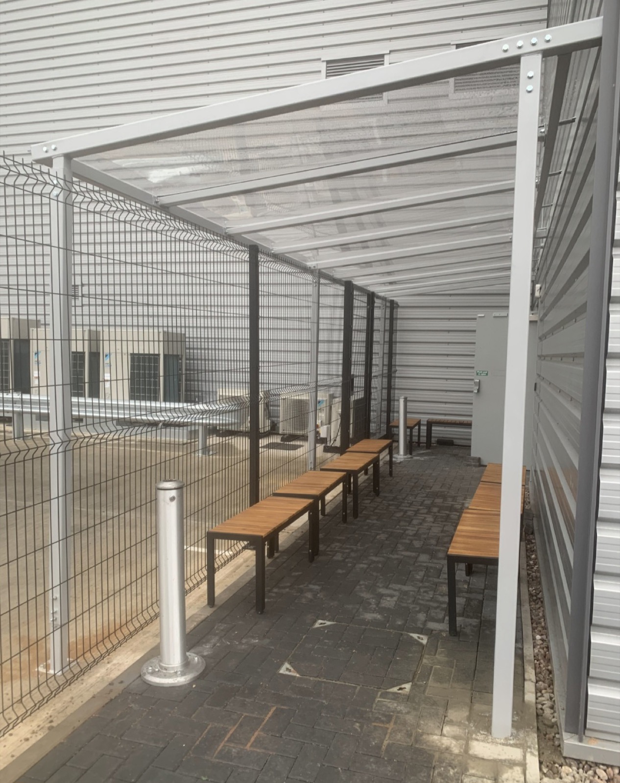 Covered walkway with wooden bench seating along a mesh fence and clad wall in Newcastle upon Tyne