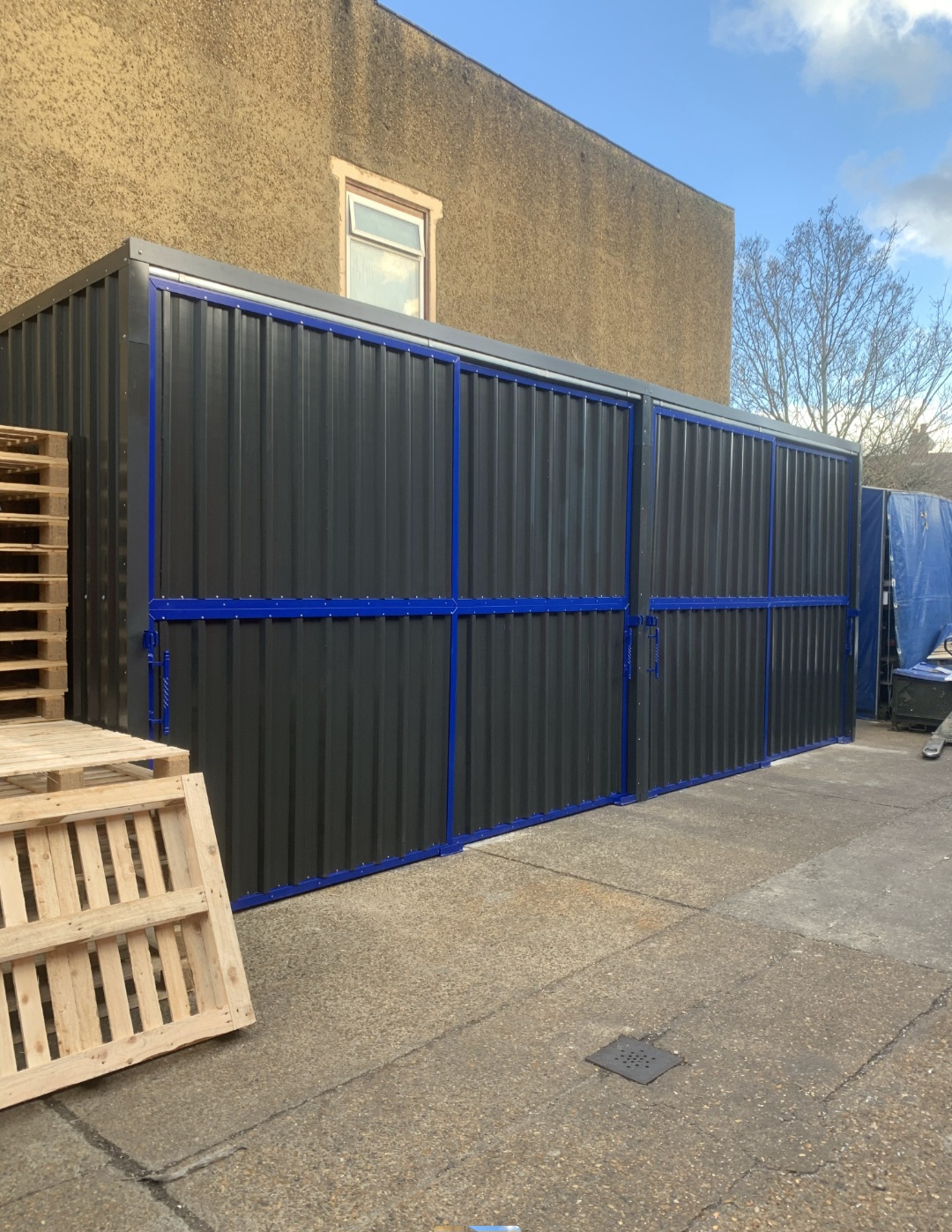 Angled view of the grey-clad storage shelter with closed blue-framed doors and wooden pallets stacked alongside