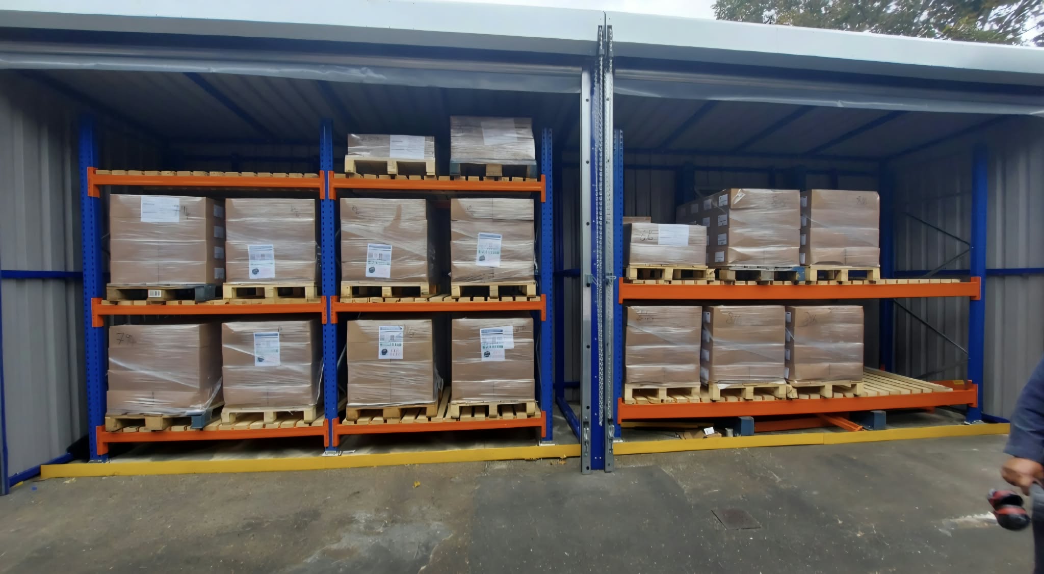 Interior pallet racking loaded with shrink-wrapped boxed stock under the storage shelter roof