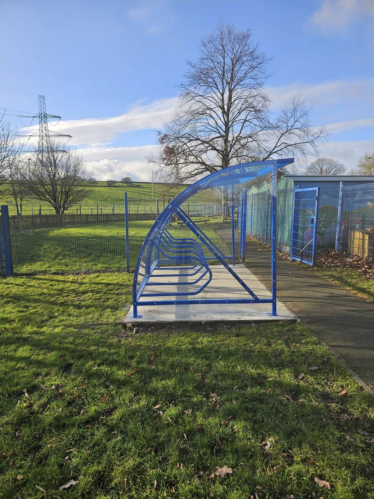 Side view of a compact blue cycle shelter with curved clear roof on a concrete base in a grassed school field