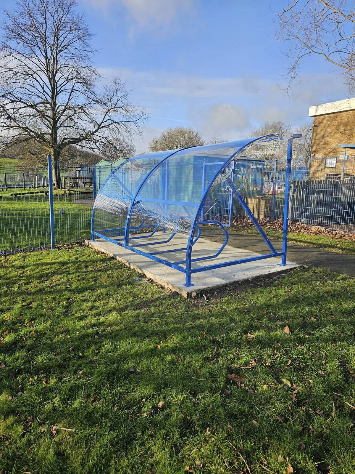 Blue cycle shelter viewed from the front, its curved transparent roof catching the light beside wire fencing