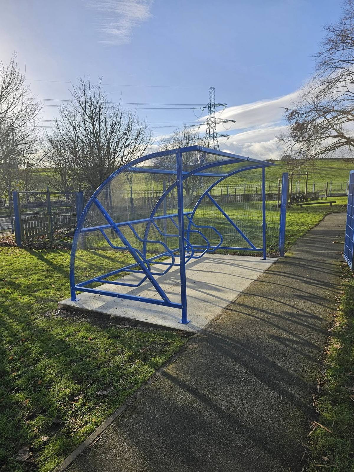 Blue cycle shelter from the open side showing the tiered rack, a pylon and playing fields behind