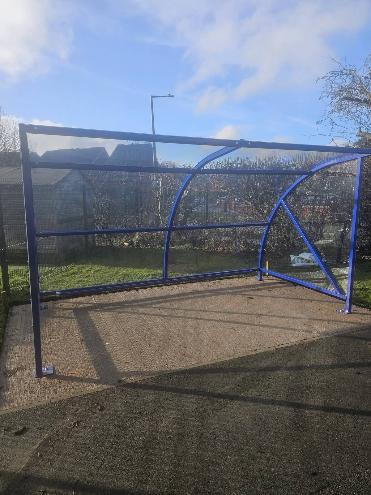 Blue curved buggy shelter frame with clear panels on a concrete base beside school grounds
